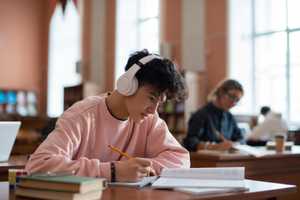 A young man with headphones sits at a desk doing coursework.
